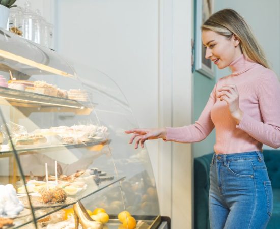 smiling-young-woman-choosing-cake-standing-front-glass-cabinet